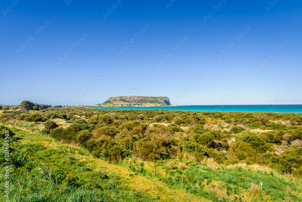 Fototapeta premium Stunning view point to big old volcanic rock mountain top called The Nut with blue turquoise water beach bay and green grass lands on warm sunny clear sky day, Stanley, North-West, Tasmania, Australia