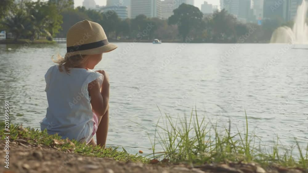 Little girl in country style hat feeding fishes in a park lake in slow motion Little girl in country style hat feeding fishes in a park lake in slow motion