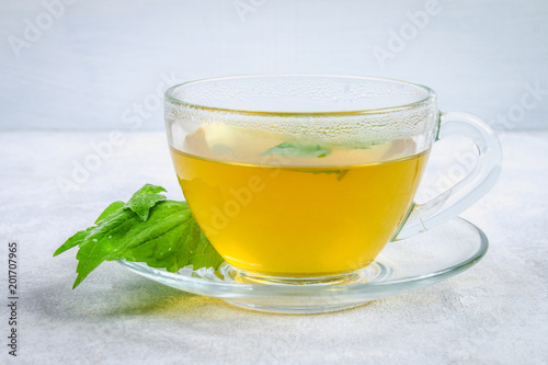 Leaves of fresh green nettle and a clear glass cup of herbal nettle tea on a gray concrete table.