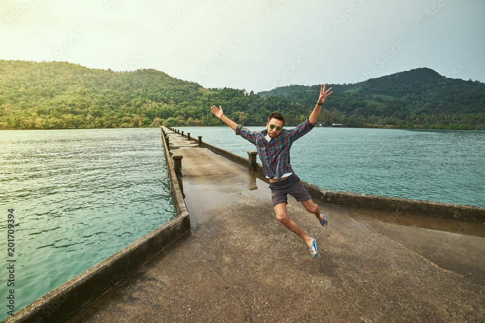 Naklejka premium Traveling boy on the pier. Pretty young man jumping on the bridge. Summer lifestyle and adventure photo