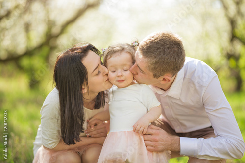 Parents kiss the baby girl in the cheeks, in the spring flowering gardens