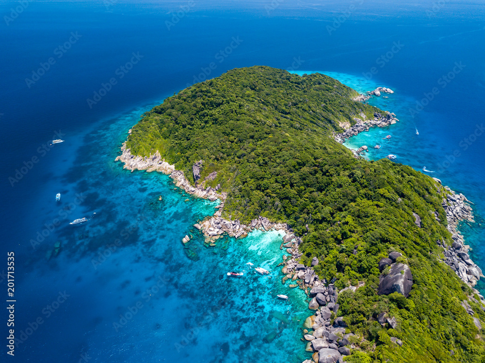 Aerial view of a boat moored in crystal clear waters over a coral reef next to a deserted, tree covered tropical island