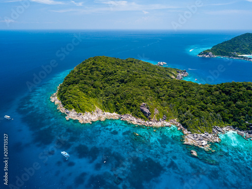 Aerial drone view of boats moored in a crystal clear ocean above a coral reef and next to a beautiful, tree covered deserted island