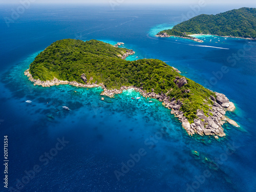 Aerial view of a boat moored in crystal clear waters over a coral reef next to a deserted, tree covered tropical island
