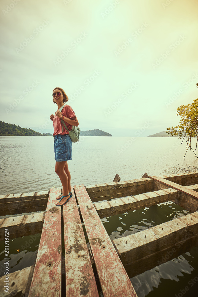 Traveling girl on the wood pier. Pretty young woman with backpack and tropical landscape. Summer lifestyle and adventure photo. Fish eye lens image Traveling girl on the wood pier. Pretty young woman with backpack and tropical landscape. Summer lifestyle and adventure photo. Fish eye lens image