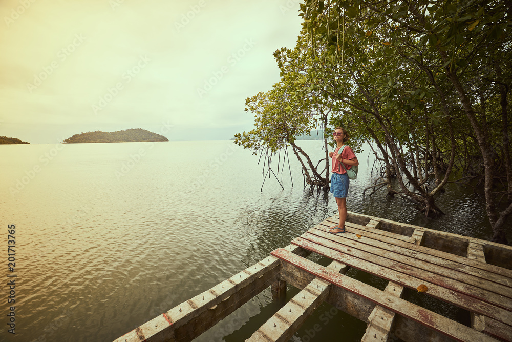 Traveling girl on the wood pier. Pretty young woman with backpack and tropical landscape. Summer lifestyle and adventure photo. Fish eye lens image