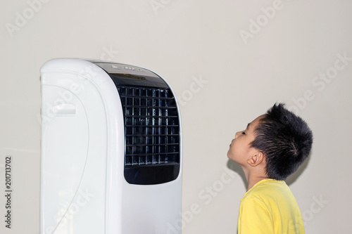 Because of the hot weather, the boy stood by the fan blowing cold air to his face.