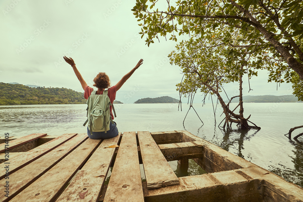 Traveling girl on the wood pier. Pretty young woman with backpack and tropical landscape. Summer lifestyle and adventure photo. Fish eye lens image