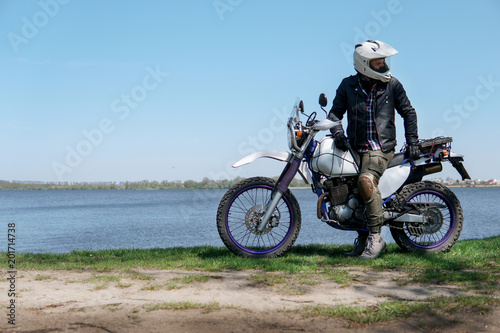 Young stylish man sit on classic retro off road track motorcycle on the beach  outdoor portrait  posing  in lather jacket and Sunglasses  travel active lifestyle concept  ocean  sea  lake  river