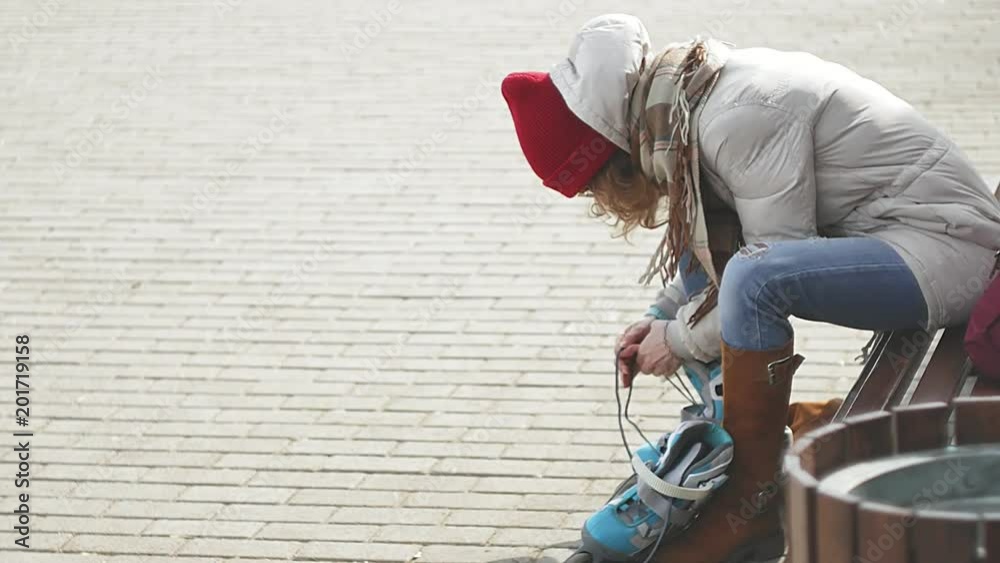 Young beautiful woman in sporty warm clothes and rollers, sitting on a wooden bench and dresses roller skates getting ready with skating Young beautiful woman in sporty warm clothes and rollers, sitting on a wooden bench and dresses roller skates getting ready with skating