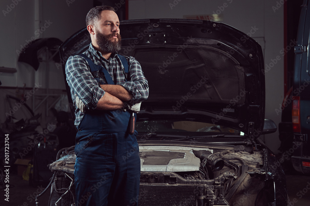 A bearded expert mechanic dressed in a uniform, standing with crossed arms against a car in the garage. A bearded expert mechanic dressed in a uniform, standing with crossed arms against a car in the garage.
