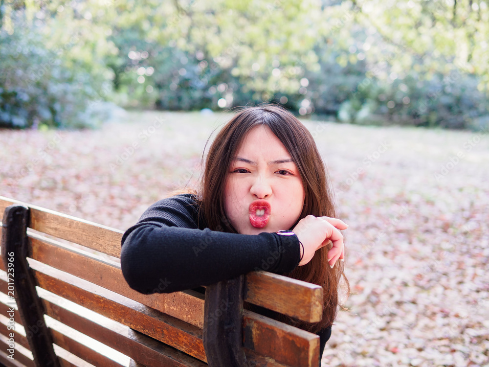 Beautiful young brunette girl sitting on bench, making face with rabbit  like teeth. Outdoor funny portrait of glamour young Chinese stylish lady.  Emotions, people, beauty and lifestyle concept. Stock Photo