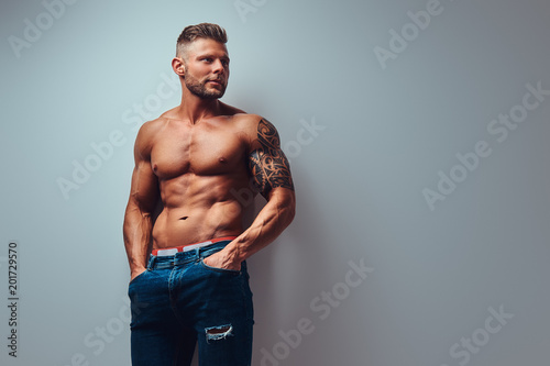 A handsome shirtless tattooed bodybuilder with stylish haircut and beard, wearing sports shorts, posing in a studio. Isolated on a gray background