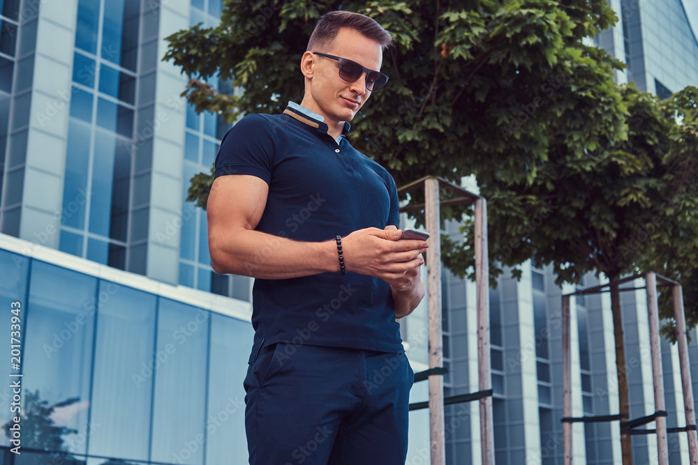 Young handsome fashionable man with a stylish haircut in sunglasses, dressed in a black t-shirt and pants, standing in the modern city against a skyscraper.