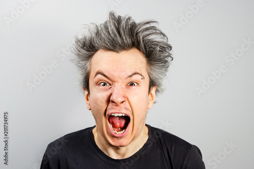 Unshorn and unshaven young guy with piercings on his face opening his mouth and screaming on gray background.