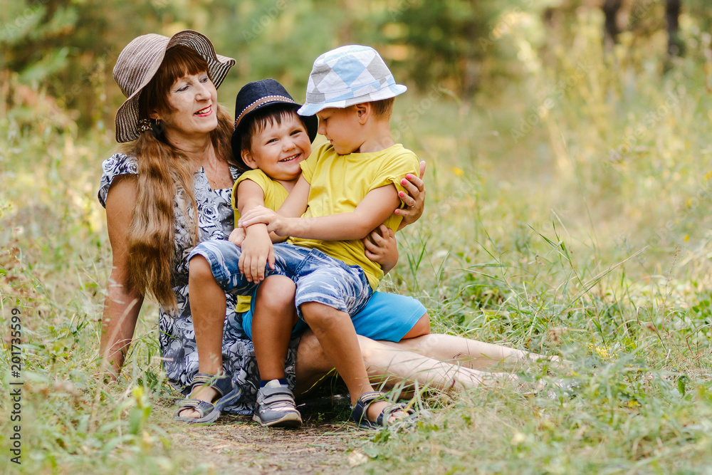 young happy grandmother with two grandchildren sitting on a forest lawn and laughing in the summer young happy grandmother with two grandchildren sitting on a forest lawn and laughing in the summer