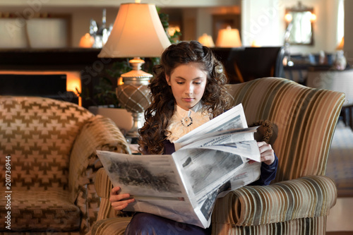marriageable girl is sitting in an old-fashioned armchair, dressed in a vintage velvet purple dress and reading a newspaper