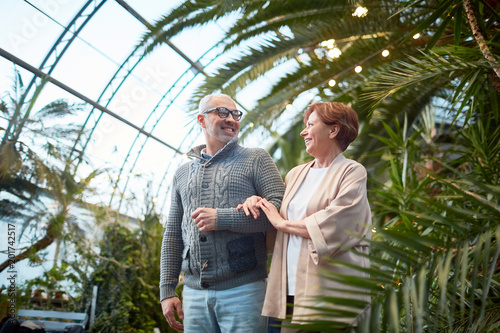 Obraz na plátně Cheerful senior man and woman having talk while walking among green plants and f