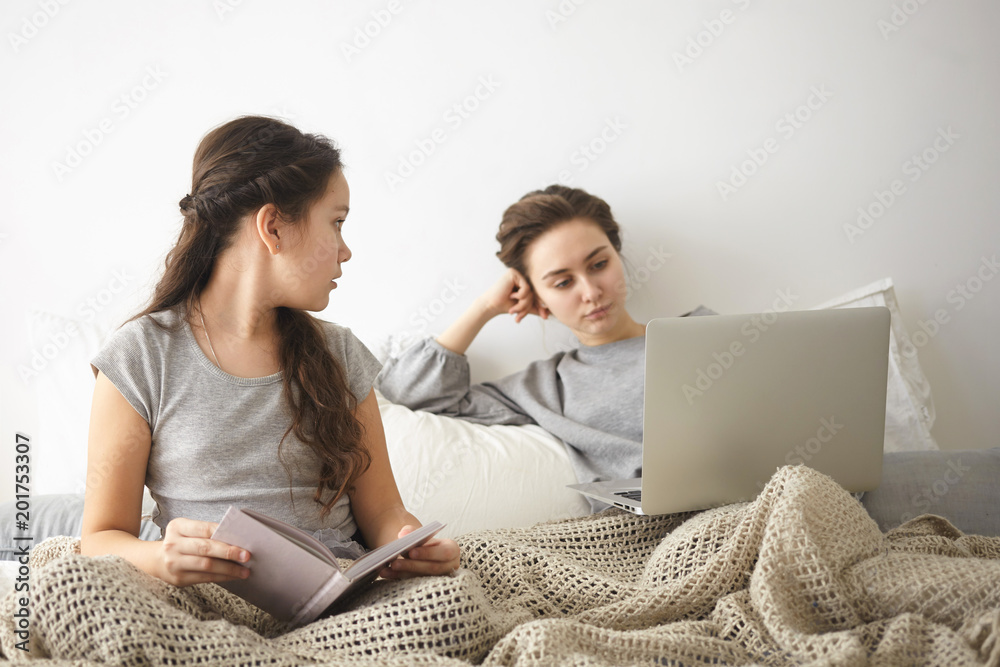 Portrait of brunette girl in gray t-shirt reading book in bed and watching her young mother surfing internet in background, having serious expression. People, leisure, technology and communication