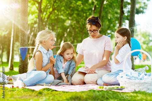 Little girls listening attentively to story or fairy-tale read by their kindergarten teacher while relaxing in park on summer day