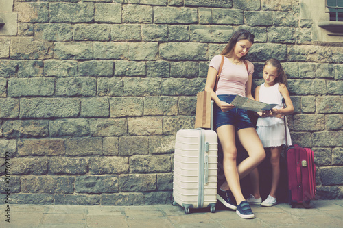 Woman and little girl traveler with suitcase leaning against stone wall