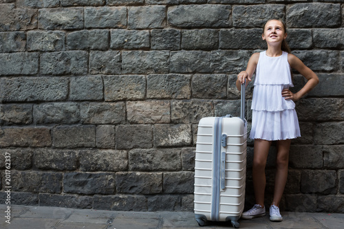 joyous little girl traveler with suitcase leaning against stone wall