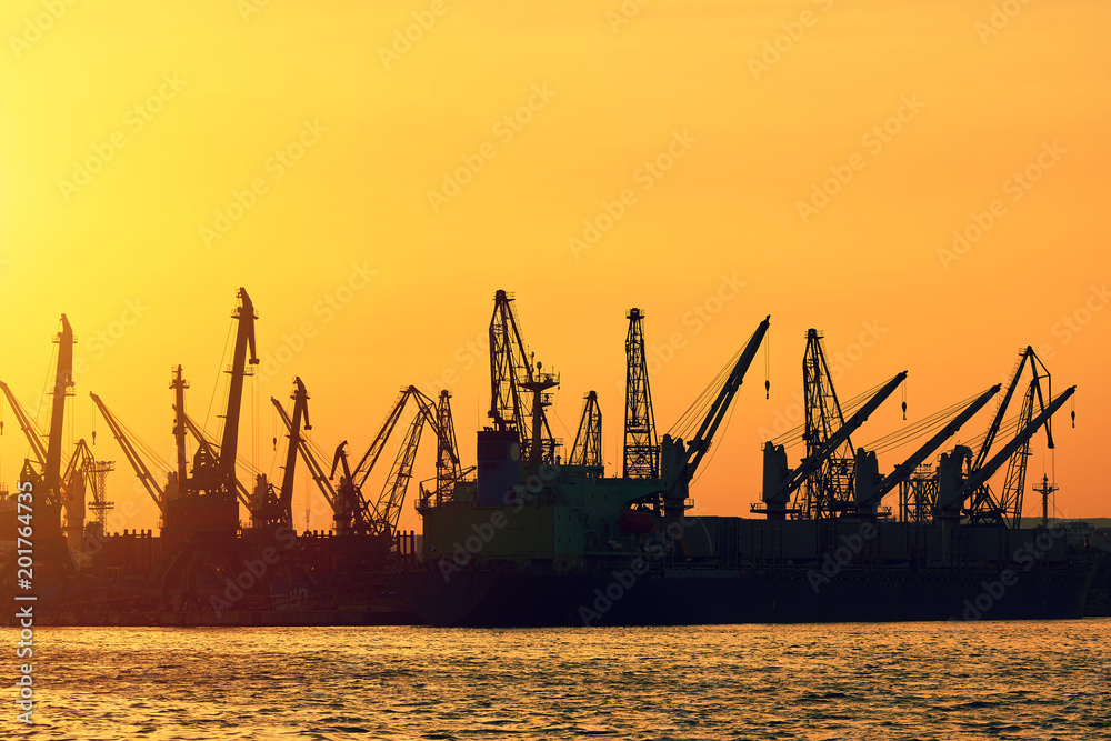 A city landscape. Bright sunset in the seaport. A sunny path on the calm surface of the sea. Large silhouettes of loading cranes. Bright abstract background ideal for any design      