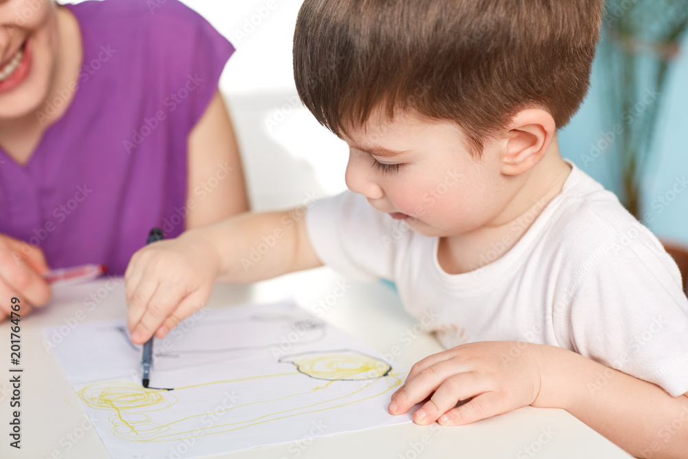 Close up shot of dark haired handsome male kid being focused at paper with picture, draws with great desire, unrecognizable female sits next to him, helps to make real masterpiece. Childhood Close up shot of dark haired handsome male kid being focused at paper with picture, draws with great desire, unrecognizable female sits next to him, helps to make real masterpiece. Childhood