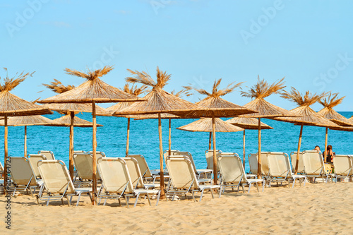 A large clean sea beach with sun loungers and thatched umbrellas for tourists on a bright sunny summer day against a blue sea and blue sky 