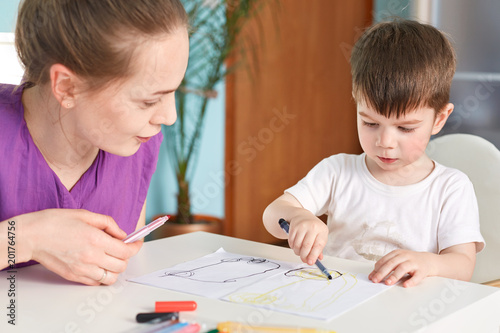 Cropped shot of concentrated small male kid holds colourful marker and makes nice picture by himself, sits near his mother who helps him and tells something interesting, cozy domestic interior