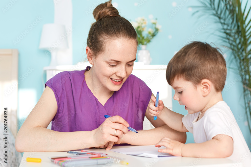 Portrait of beautiful female with hair knot, dressed in casual purple t shirt, draws together with her little son, tries to develop his mind and prepare for school. Children and creativity concept