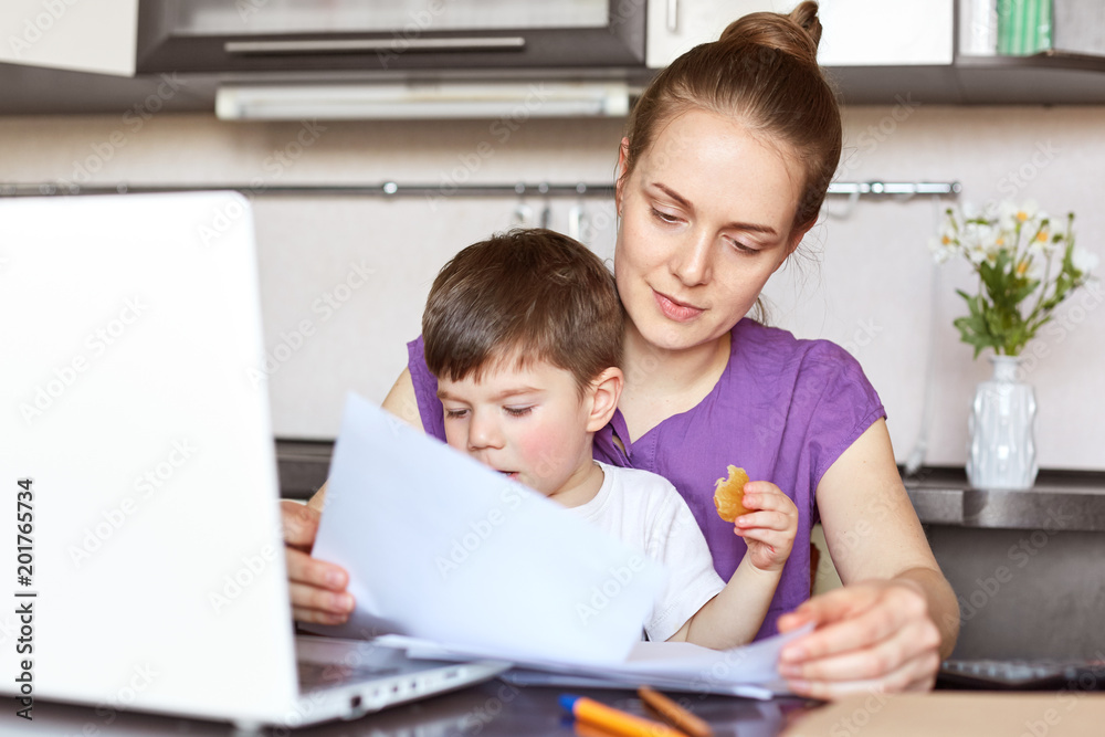 Mother freelancer on maternity leave works with documentation, makes business report on portable laptop computer, holds son who interrupts her, sit together against kitchen interior. Family concept Mother freelancer on maternity leave works with documentation, makes business report on portable laptop computer, holds son who interrupts her, sit together against kitchen interior. Family concept