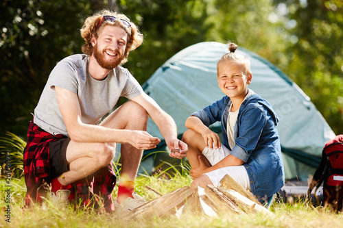 Happy young man with box of matches and his son looking at camera with pile of woods near by © pressmaster