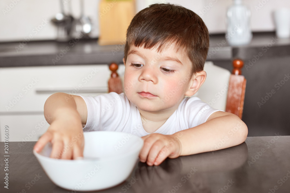Attractive boy with pleasant appearance, sits at kitchen table, looks in white bowl, waits for dinner, being hungry after playing with friends, being at home. Children and nutrition concept. Attractive boy with pleasant appearance, sits at kitchen table, looks in white bowl, waits for dinner, being hungry after playing with friends, being at home. Children and nutrition concept.