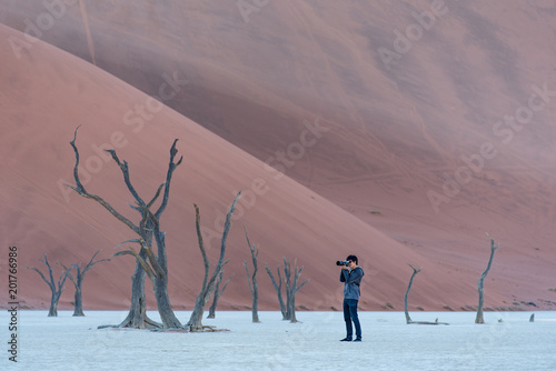 Young male photographer and traveler taking photo of dead tree in deadvlei (Sossusvlei), famous natural landmark in Namib desert of Namibia, Africa. Travel photography concept