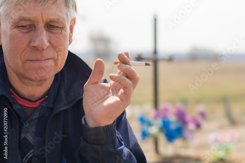 a gray-haired village man a widower sadly sits near a fresh grave and smokes a cigarette