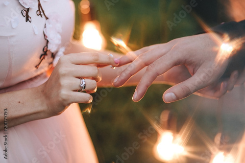 Wedding ceremony. Bride in a pink dress is wearing an engagement ring to the groom on the background of a bokeh of light bulbs. On her hand is a gold ring with a diamond close-up. photo