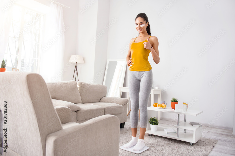 Content with results. Beautiful young woman standing on the scales and weighing herself while showing thumbs up and being satisfied with the result Content with results. Beautiful young woman standing on the scales and weighing herself while showing thumbs up and being satisfied with the result
