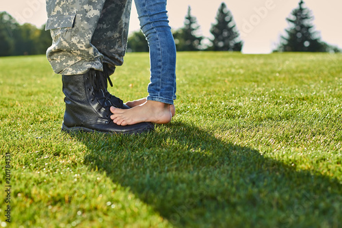 Close up feet of soldier and daughter. Leather military boots and naked feet on the meadow.