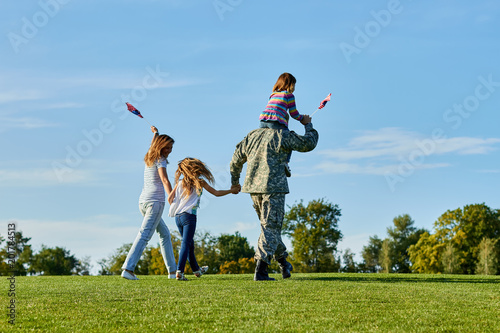Soldier is walking with his family in the grass, back view. Patriotic family waving flags outdoors on the park meadow grass.