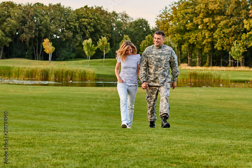 Soldier and his girlfriend are walking on the grass. Holding hands, happy couple walking after years of separation on green lawn.