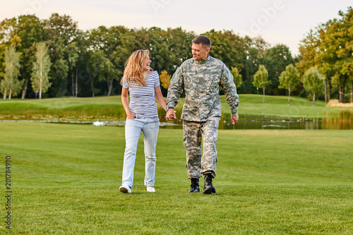 Soldier walking with woman, holding hands. Happy couple on the grass.
