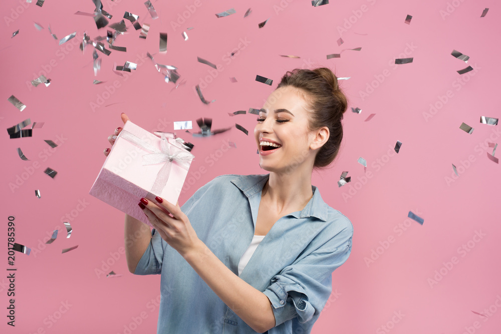 Happiness. Cheerful cute girl is demonstrating gift box being surrounded by confetti. She is expressing gladness. Pink background - obrazy, fototapety, plakaty 