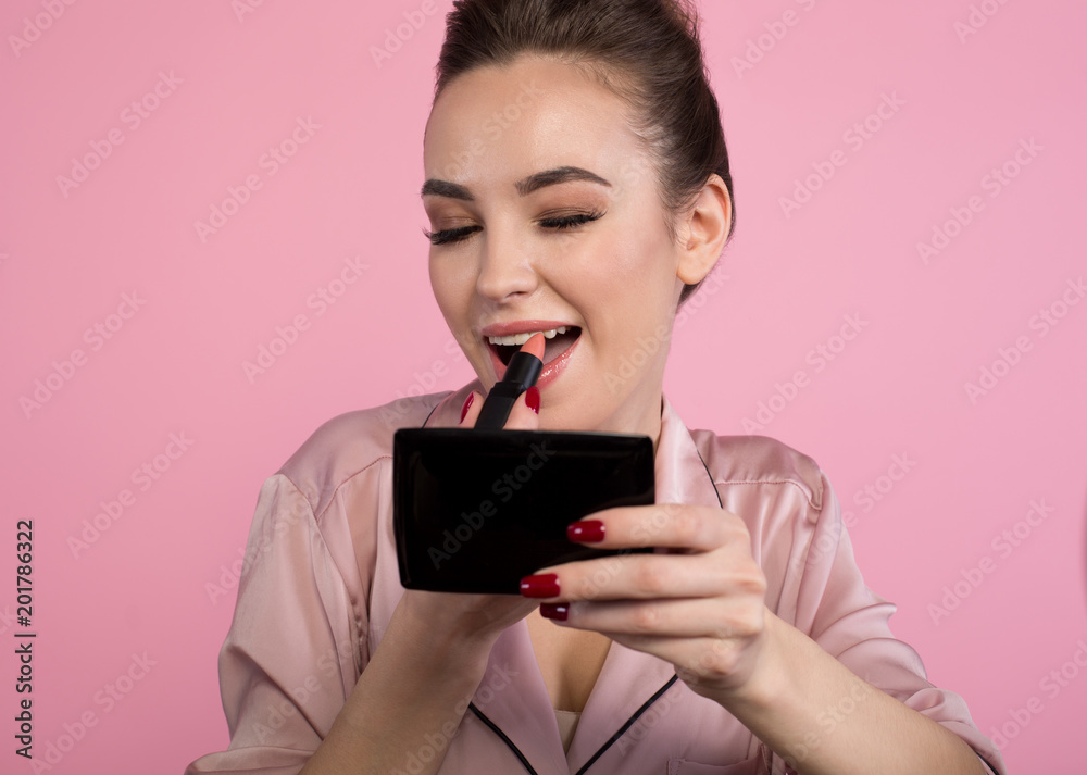 Playful mood. Portrait of optimistic girl is sitting and applying lipstick on her lips with smile. She is looking at small mirror in her hand. Isolated on pink background Playful mood. Portrait of optimistic girl is sitting and applying lipstick on her lips with smile. She is looking at small mirror in her hand. Isolated on pink background