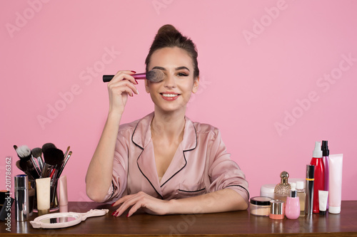 Portrait of optimistic young female cosmetologist is sitting at dressing table with cosmetics items and holding brush Fototapeta