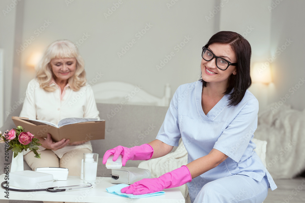 Obraz premium Weekly cleaning. Elder woman reading book while cheerful caregiver making house clean
