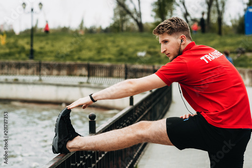 Photos Handsome sporty guy is streching in a green beautiful park and is getting ready for jogging while is listening music