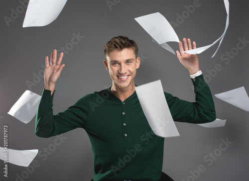 Waist up portrait of satisfied guy tossing away documents in the air while sitting. He is staring at camera with joy. Isolated on background photo