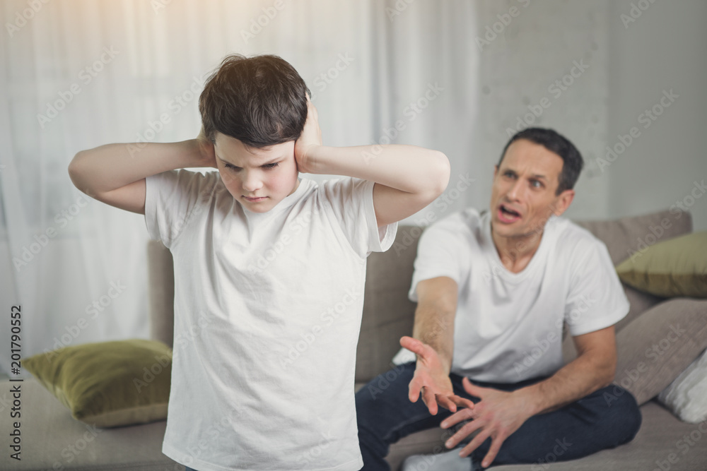 I do not want to hear you. Sad boy is closing his ears while his dad is shouting at him. Angry man is sitting on couch on background I do not want to hear you. Sad boy is closing his ears while his dad is shouting at him. Angry man is sitting on couch on background