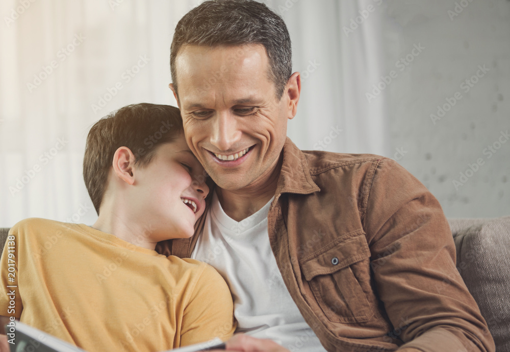 Portrait of happy father reading fairy tale for his lovely son. Boy is leaning head on male shoulder and laughing